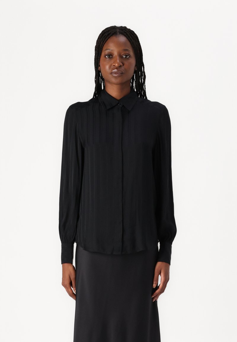 Black long-sleeve shirt with a button-down collar, featuring vertical pleats and a smooth texture. Worn with a matching dark skirt.