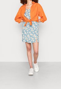 Oranje overhemd geknoopt in de taille over een blauwe jurk met witte madeliefjes en een zijsplit, gecombineerd met witte sneakers.