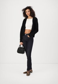 Zwarte cardigan over een witte cropped top, donkere jeans, zwarte gestoffeerde handtas en loafers met luipaardprint. Krullend haar en een neutrale achtergrond.