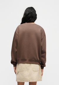 Καφέ φούτερ με χαμηλωμένους ώμους και ριμπ μανσέτες, συνδυασμένο με μπεζ φούστα με δύο πίσω τσέπες. Υφή υφάσματος, άνετη εφαρμογή.