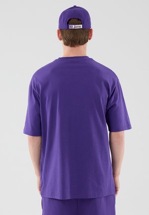 Osoba nosząca fioletową czapkę Lakers i pasującą oversize koszulkę, pokazana od tyłu na jednolitym szarym tle.