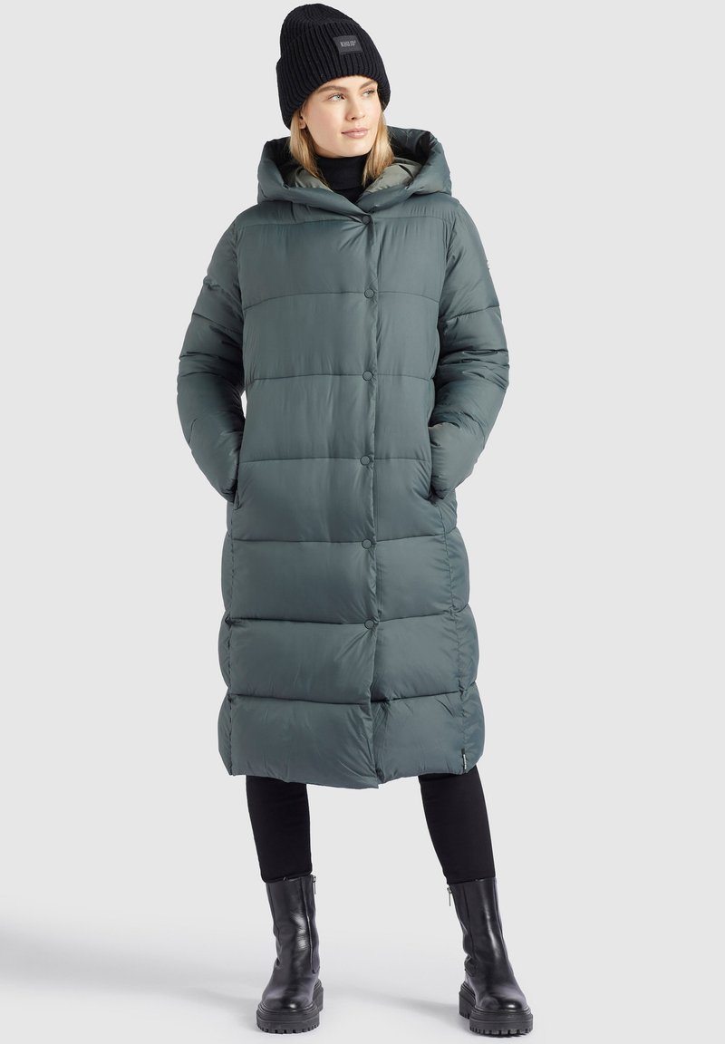 Langer, gesteppter, grüner Puffermantel mit Kapuze, der über einen Knopfverschluss und seitliche Taschen verfügt. Getragen über einem schwarzen Oberteil und einer Hose, kombiniert mit schwarzen Stiefeln.