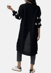 Long cardigan noir en tricot avec des accents d'étoiles sur les manches, une fente dans le dos et une finition texturée, associé à un jean gris et des baskets.