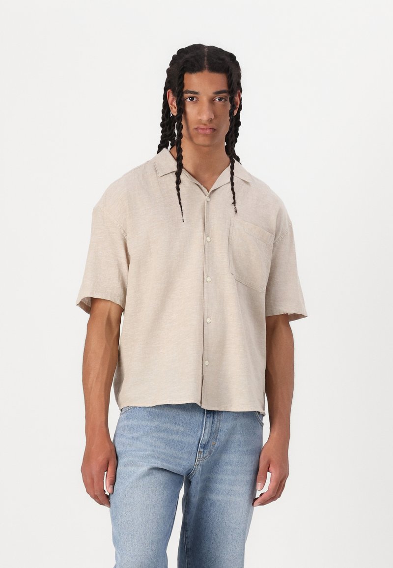 Chemise en lin beige à manches courtes avec une fermeture à boutons, un col pointu et une poche poitrine. Elle présente une coupe décontractée et un tissu texturé.