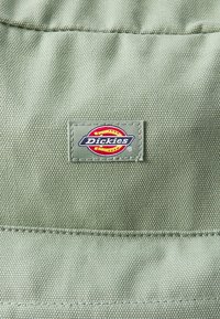Tela texturizada verde con una etiqueta de logo de Dickies cosida que presenta un diseño circular en rojo, amarillo y azul. Costuras simples y resistentes.