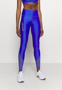 Korkeavyötäröiset siniset legginsit, joissa on kiiltävä pinta. Niissä on tummansiniset kontrastipaneelit ja vihreä "Under Armour" -logo sivussa.