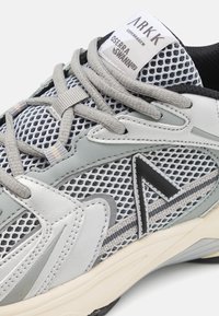 Sapatilha cinza claro com parte superior em malha, detalhes em couro e detalhes pretos. Apresenta atacadores, colarinho acolchoado e sola texturizada.