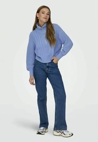 Kvinde står med hånden i lommen iført en lysblå kabelstriktrøje, mørkeblå jeans og hvide løbesko på ensfarvet baggrund.