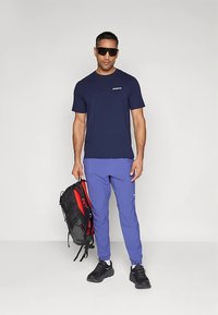 T-shirt azul-marinho, calças atléticas roxas, ténis pretos e óculos de sol. Mochila preta com detalhes em vermelho. Design simples, ajuste casual.