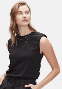 Zwarte mouwloze top in een zachte stof, met een ronde halslijn. Het model draagt gelaagde gouden kettingen, tegen een effen witte achtergrond.