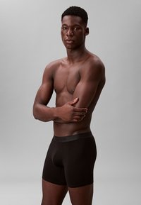 Czarne bokserki wykonane z teksturowanego materiału w pasie. Model o szczupłej sylwetce, z założonymi na siebie ramionami, stojący na neutralnym szarym tle.