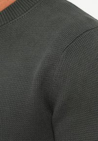 Pull en tricot vert foncé avec un col côtelé et un tissu texturé, mettant en avant la durabilité et la chaleur. Gros plan sur la zone de l'épaule.