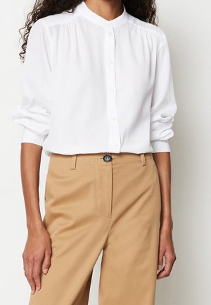 Donna che indossa una camicia bianca con bottoni e maniche lunghe e pantaloni beige a vita alta con un bottone nero.