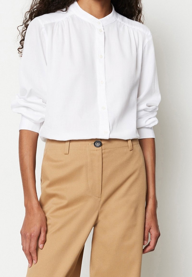 Femme portant une chemise blanche boutonnée à manches longues et un pantalon beige taille haute avec un bouton noir.