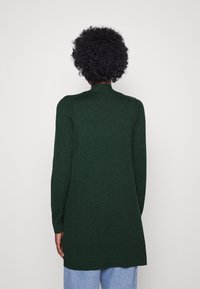 Długi kardigan z długim rękawem w ciemnozielonym kolorze, wykonany z miękkiego materiału. Posiada wysoki dekolt i ściągacz na dolnym brzegu, charakteryzuje się luźnym krojem oraz długością do połowy uda.