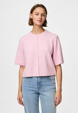 Cardigan corto rosa a coste con maniche corte, scollatura rotonda e bottoni frontali, abbinato a jeans azzurri chiari. Tessuto morbido, vestibilità casual.