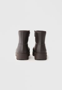 Botas de tobillo de goma marrón con una textura suave, cremalleras laterales y una suela gruesa y texturizada, con sutiles líneas verticales en los lados.