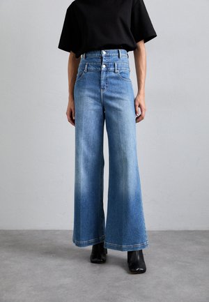 Világoskék, magas derekú, bő szárú farmernadrág sima denim textúrával, két elülső gombbal és rojtos szegéllyel, fekete bokapapuccsal párosítva.