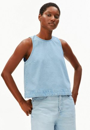 Femme portant un haut en denim bleu clair sans manches et un pantalon denim assorti taille haute, debout les mains sur les hanches sur fond blanc.