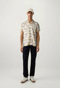 Kortærmet skjorte med en cremefarvet baggrund, prydet med et blomstermotiv og dyreprint i beige og brun, parret med mørke jeans og sneakers.