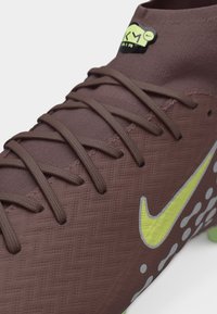 Κοντινό πλάνο καφέ αθλητικού παπουτσιού με υφασμάτινη υφή, κορδόνια στο ίδιο χρώμα, φωτεινό κίτρινο λογότυπο Nike και ορατά πράσινα καρφιά στη σόλα.