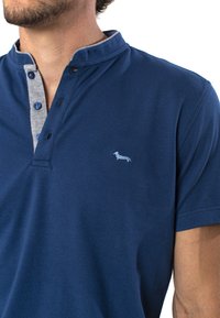 Námořnická modrá polo košile s šedým límečkem na knoflíky, který má modré knoflíky a malé vyšívané logo psa na hrudi. Bavlněný materiál.