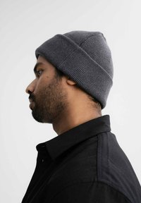 Grijze gebreide beanie met een geribde textuur. Opgerold op het hoofd van een persoon, zijprofiel zichtbaar tegen een neutrale achtergrond.