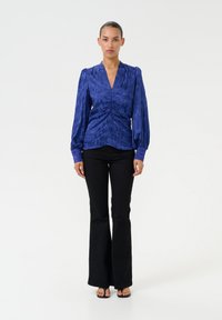 Blue textured blouse with a v-neck and gathered detailing at the waist, paired with flared black trousers and flat sandals.