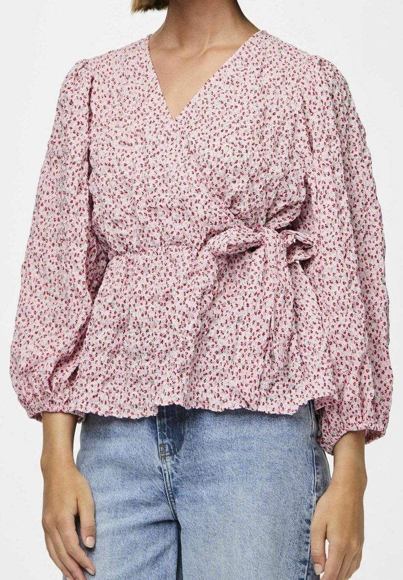 Top floreale rosa avvolgente con maniche a palloncino, tessuto texturizzato e vita legata. Abbinato a jeans denim azzurri.