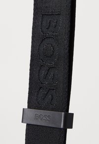 Černý textilní pásek s embosovaným logem "BOSS" a matnou černou obdélníkovou sponou. Materiál má strukturovaný tkaný vzor.
