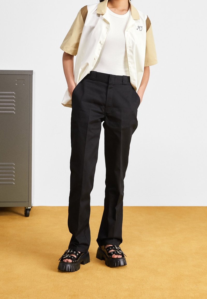 Personne portant un pantalon noir sur mesure, un haut blanc sans manches, une chemise beige et blanche à manches courtes et des sandales noires épaisses à brides, debout à l’intérieur.