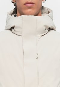Giacca beige chiaro con colletto alto e cappuccio regolabile, dotata di una zip frontale e di una texture di tessuto liscia e opaca.