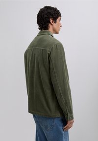 Hombre de perfil mirando a la derecha, vistiendo una chaqueta de pana verde oliva y jeans azules, sobre un fondo gris claro liso.