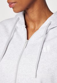 Ljusgrå zip-up hoodie av mjukt tyg med texturerad finish. Har en justerbar huva och minimalistisk logotypdetalj på bröstet.