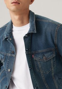 Donkerblauwe denimjack met een gestructureerd ontwerp, voorzien van metalen knopen, gestikte accenten en een borstzak aan de voorkant. Daaronder een wit T-shirt.