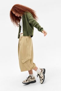 Cardigan verde, top cortado blanco, pantalones cortos cargo caqui con bolsillos laterales y zapatillas de patrón con suelas negras, sobre un fondo blanco.