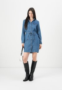 Denim skjortekjole med lange ermer, frontknapper, to brystlommer, justerbart midjebånd og lengde til midten av låret, kombinert med knelang svarte støvler.
