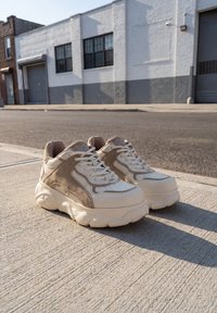 Stevige beige en witte sneakers naast elkaar geplaatst op een betonnen stoep met een stedelijk gebouw op de achtergrond.