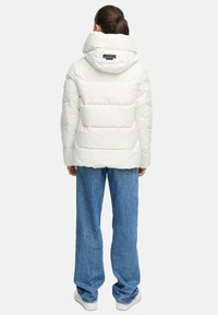 Vitt pufferjacka med huva, quiltat mönster och en märkespatch på ryggen, i kombination med löst sittande blå denimbyxor och sneakers.