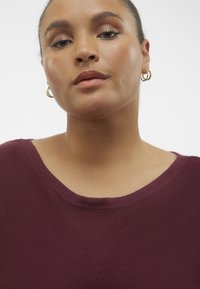 Burgundská žebrovaná pletená halenka s širokým kulatým výstřihem. Modelka má na sobě malé zlaté kruhové náušnice. Hladká textura je viditelná na látce.