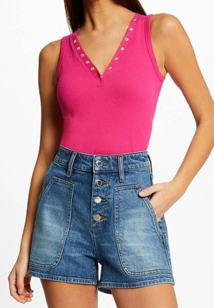 Vrouw draagt een felroze mouwloos geribbeld topje met zilveren studs bij de halslijn en hooggetailleerde blauwe denim shorts met grote voorzakken.