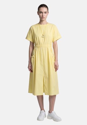 Woman wearing a yellow knee-length dress with short sleeves, white sneakers, and a round pendant necklace, standing against a plain background.