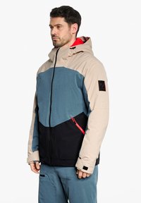 Farbblockierte Jacke mit beigen, blauen und schwarzen Paneelen, rotem Innenfutter, Reißverschlusstasche und Logopatch am Ärmeln.