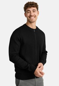 Cardigan negro con cremallera, puños y dobladillo acanalados, textura de punto, cuello redondo, con un diseño sutil. La modelo posa con las manos juntas.