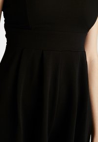 Robe noire avec un bodice ajusté, une taille large et une jupe plissée fluide, fabriquée en tissu lisse avec une texture mate.