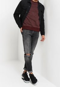 Schwarze Jeansjacke, dunkelroter strukturierter Pullover, abgenutzte graue Jeans und schwarze Turnschuhe mit transparenten Sohlen. Keine weiteren Details oder Hintergrund.