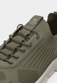 Chaussure de sport tricotée vert olive avec lacets élastiques, côtés texturés et semelle blanche, marquée "MOP" sur la languette.