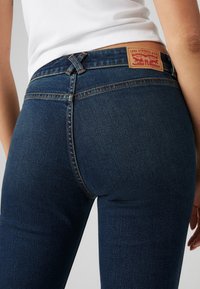 Jean en denim bleu marine avec une taille haute, des surpiqûres discrètes et un patch arrière en cuir classique avec un logo embossé. Texture lisse.