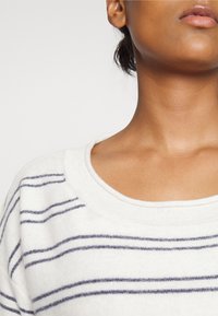 Vit tröja med marinblå horisontella ränder, bred halsringning och mjukt, texturerat tyg. Nära bild av kragen och nackområdet.