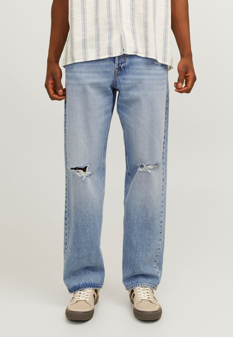 Jean en denim bleu clair avec une coupe décontractée, présentant des fentes déchirées aux genoux et des détails de couture. Portés avec des baskets beiges.